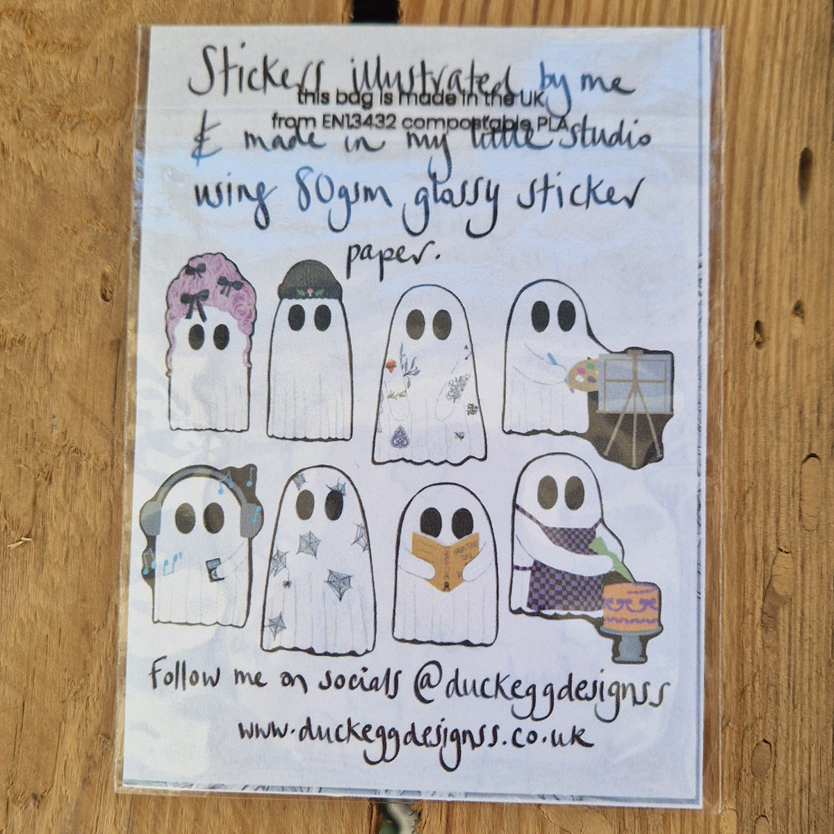 A sticker set lies on a wooden plank background face down, showing the eight different ghost designs inside as well as information about the stickers written in black handwriting.
