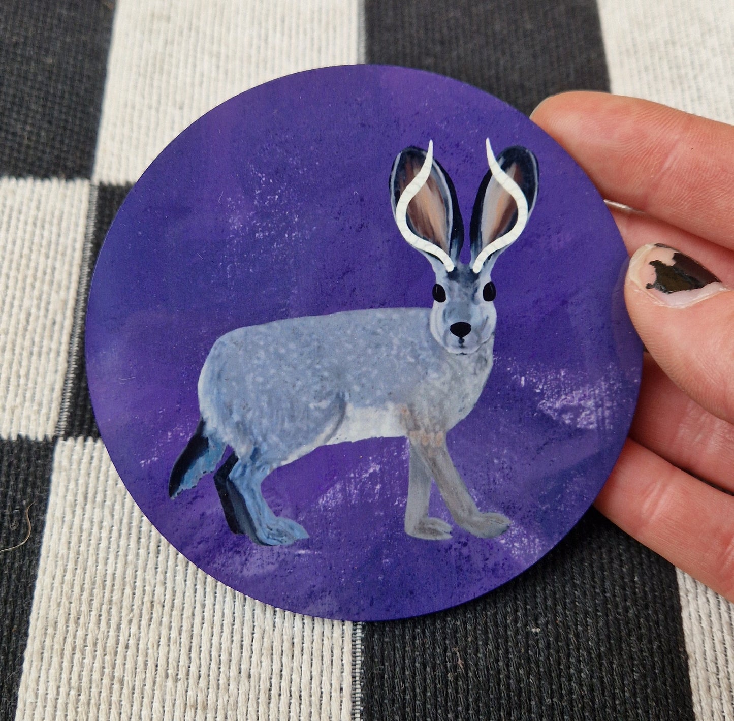 A round mottled rich purple coaster with a grey and black jackalope on it. The coaster is held in front of a checkered white and black fabric by a white hand.
