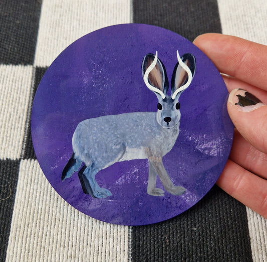 A round mottled rich purple coaster with a grey and black jackalope on it. The coaster is held in front of a checkered white and black fabric by a white hand.