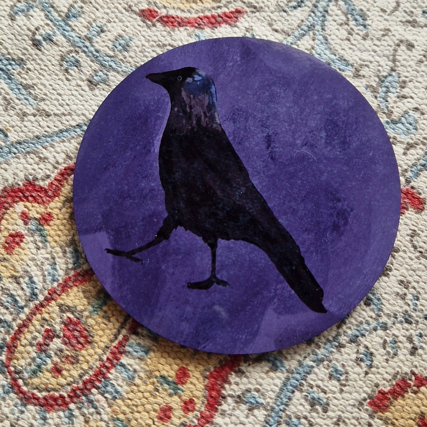 A round purple coaster with a black jackdaw design sits on a patterned white fabric background.