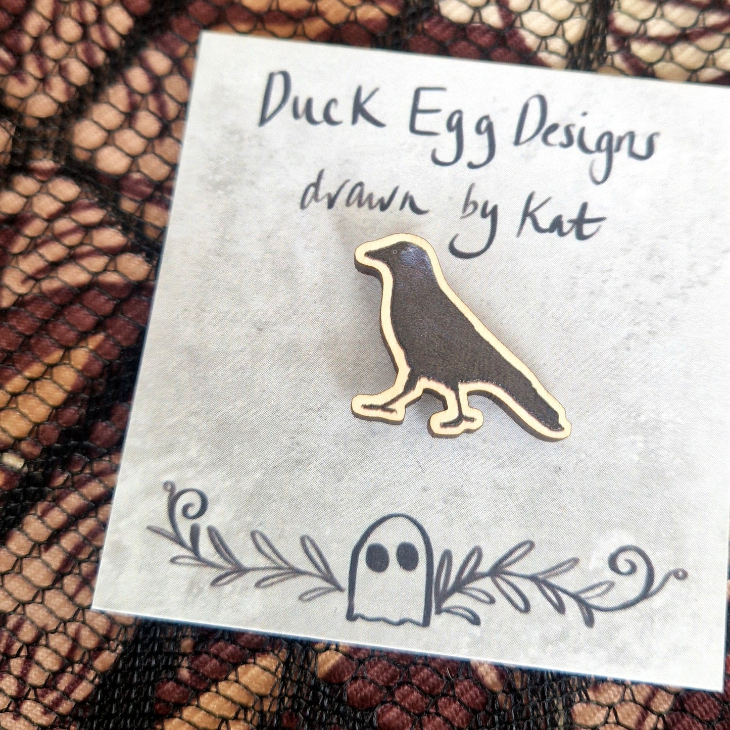 A black jackdaw pin badge sits on a grey backing card with the words 'Duck Egg Designs drawn by Kat' written in black across the top. Behind the backing card is a black lace fabric on top of a brown patterned background.