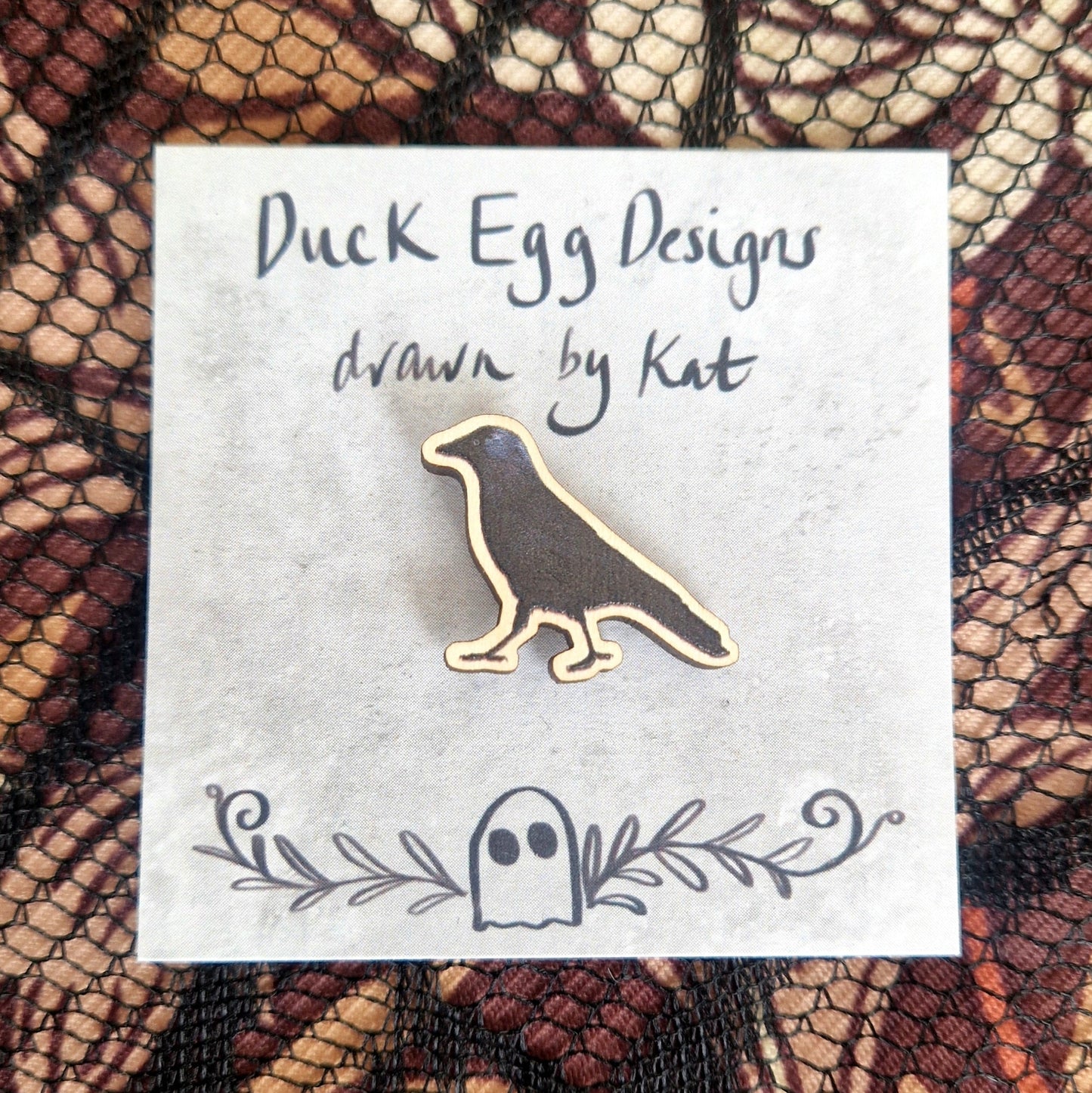 A black jackdaw pin badge sits on a grey backing card with the words 'Duck Egg Designs drawn by Kat' written in black across the top. Behind the backing card is a black lace fabric on top of a brown patterned background.