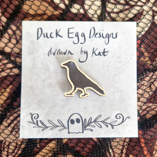 A black jackdaw pin badge sits on a grey backing card with the words 'Duck Egg Designs drawn by Kat' written in black across the top. Behind the backing card is a black lace fabric on top of a brown patterned background.