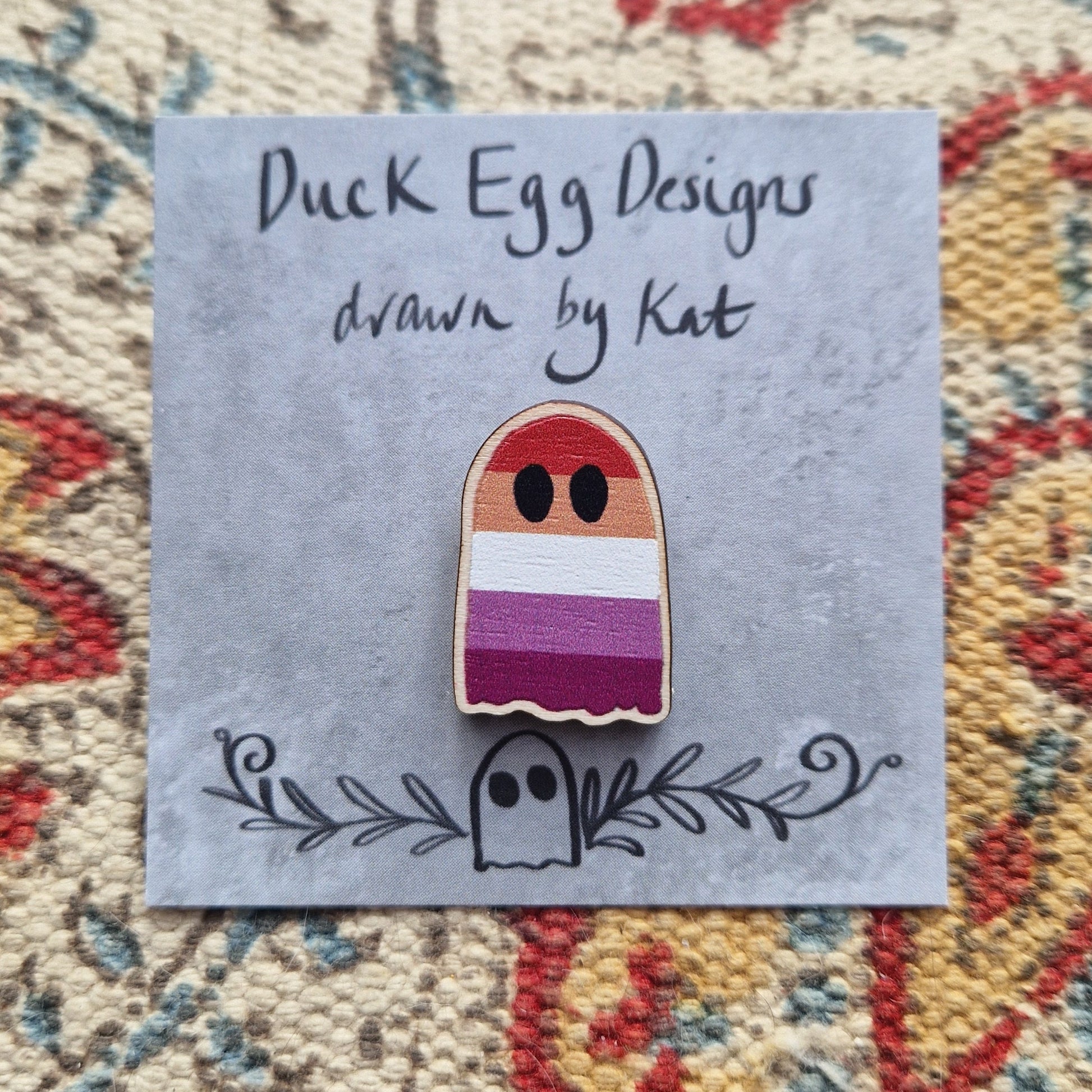 A ghost pin with the stripes of the lesbian flag design running across it sits on a grey square backing card with the words 'Duck Egg Designs drawn by Kat' across the top. Behind the backing card is a patterned fabric background.