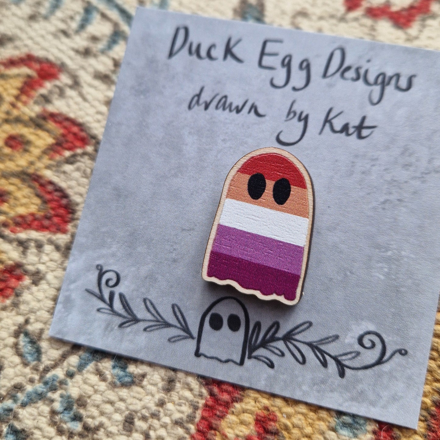 A ghost pin with the stripes of the lesbian flag design running across it sits on a grey square backing card with the words 'Duck Egg Designs drawn by Kat' across the top. Behind the backing card is a patterned fabric background.