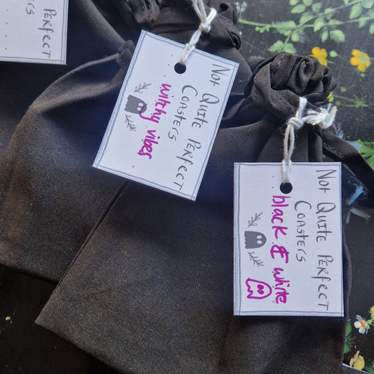 Three black fabric pouches lying next to each other on a black wooden table with flowers painted on it. The pouches have white labels that say 'Not Quite Perfect Coasters' in black handwriting with purple writing that says the theme of the coasters inside.