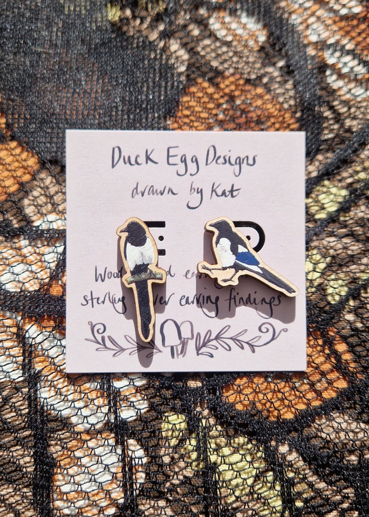 Mismatched magpie earrings sit on a light purple backing card with the words 'Duck Egg Designs drawn by Kat' in black handwriting. Behind the backing card is a black lace and floral fabric background.