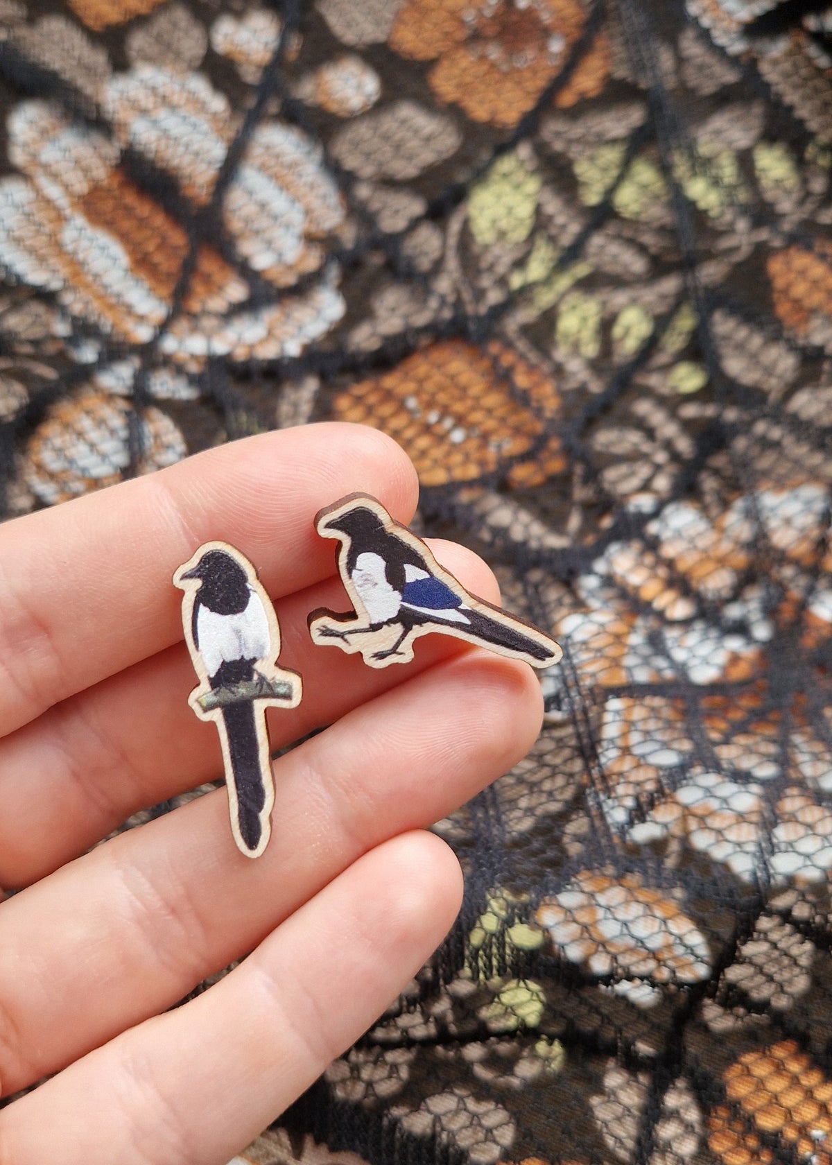 A pair of magpie earrings sit on a white hand above a black lace and floral fabric background. 