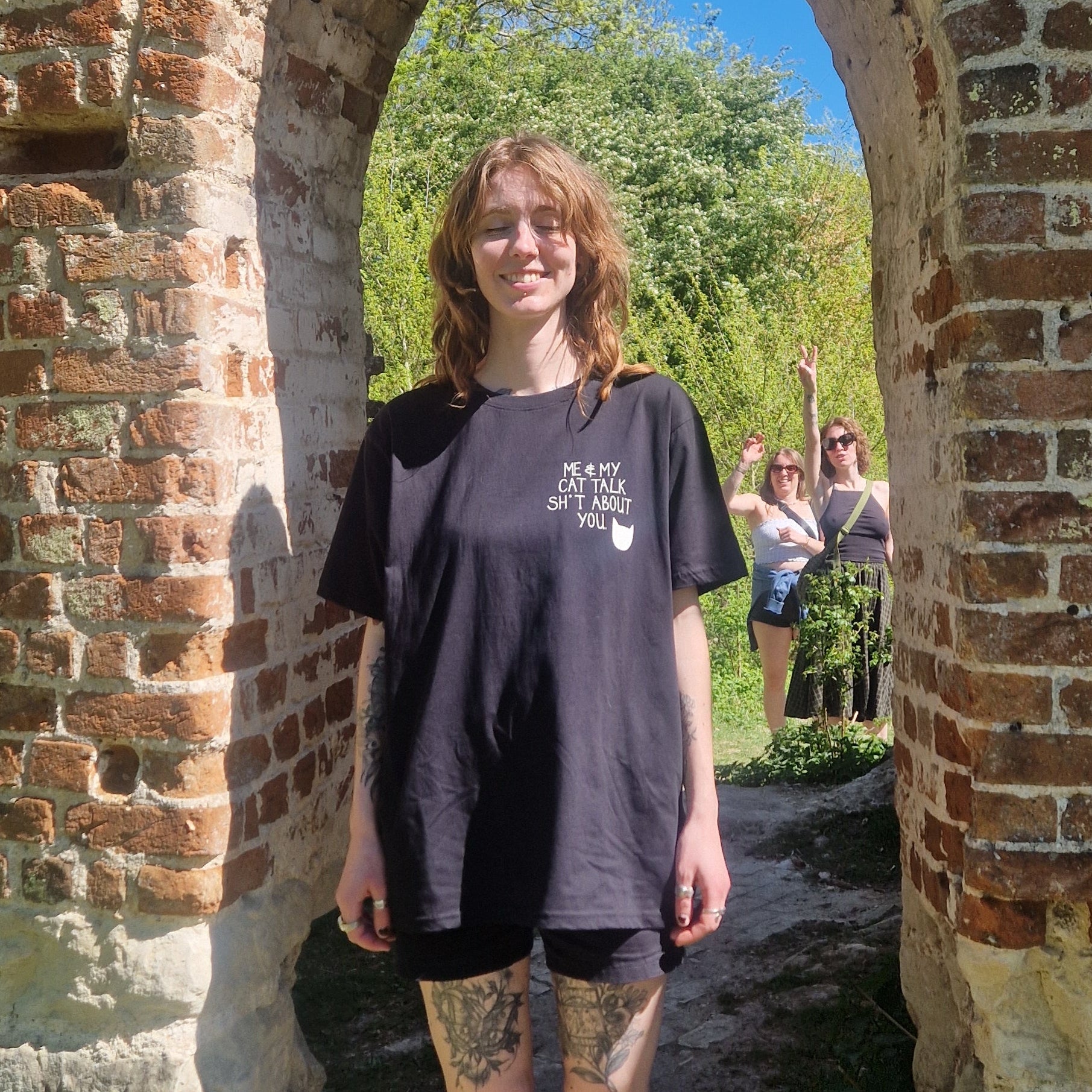 A white woman in an oversized black tshirt with a small print on the right of the shirt that reads 'ME & MY CAT TALK SH*T ABOUT YOU.' next to a white cat head silhoette. The woman stands in a brick archway with trees and people behind her. 
