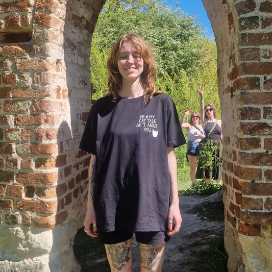 A white woman in an oversized black tshirt with a small print on the right of the shirt that reads 'ME & MY CAT TALK SH*T ABOUT YOU.' next to a white cat head silhoette. The woman stands in a brick archway with trees and people behind her. 