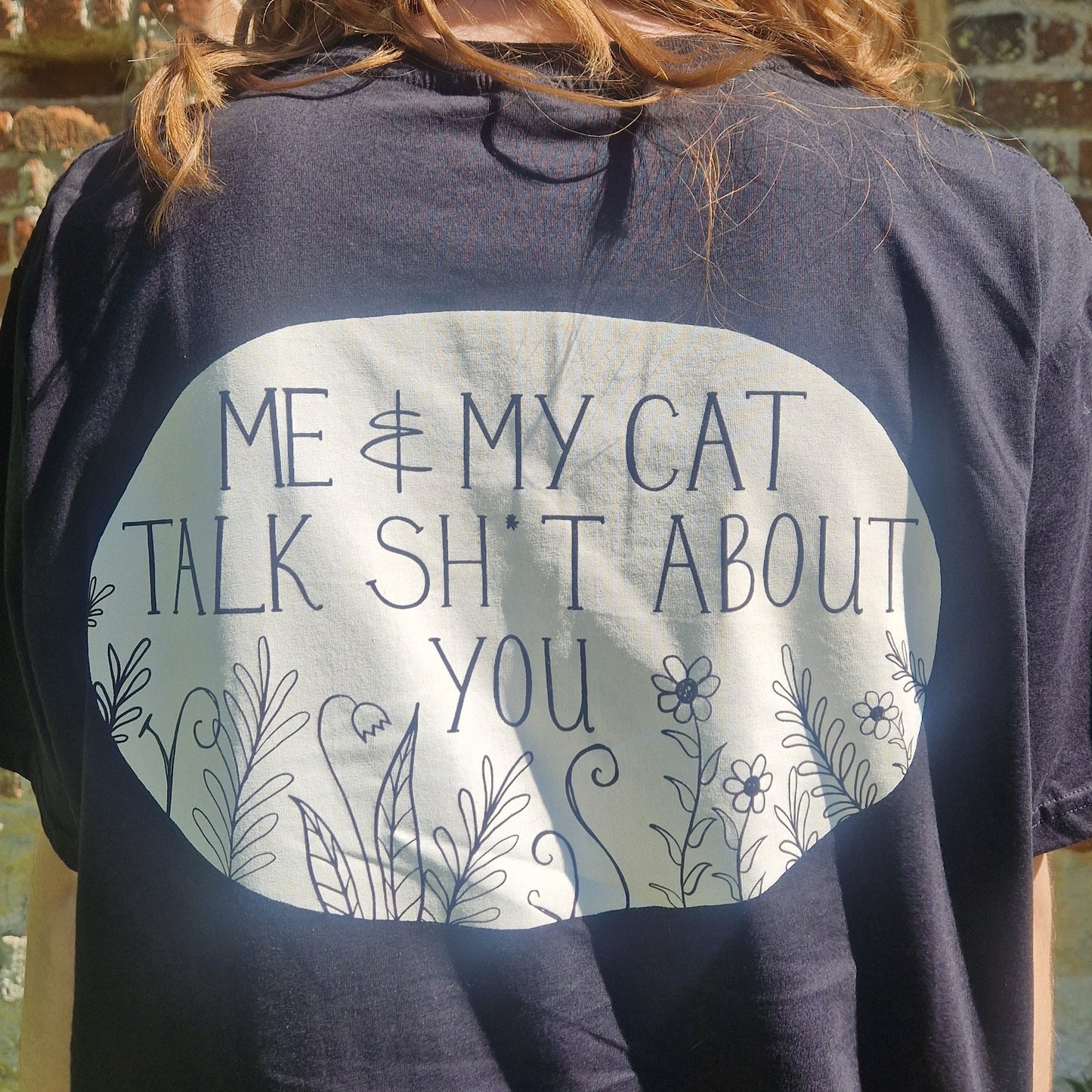 A close up of the back print of the black t-shirt showing the white oval with black writing that reads 'ME & MY CAT TALK SH*T ABOUT YOU' above black flowers and leafy fronds.