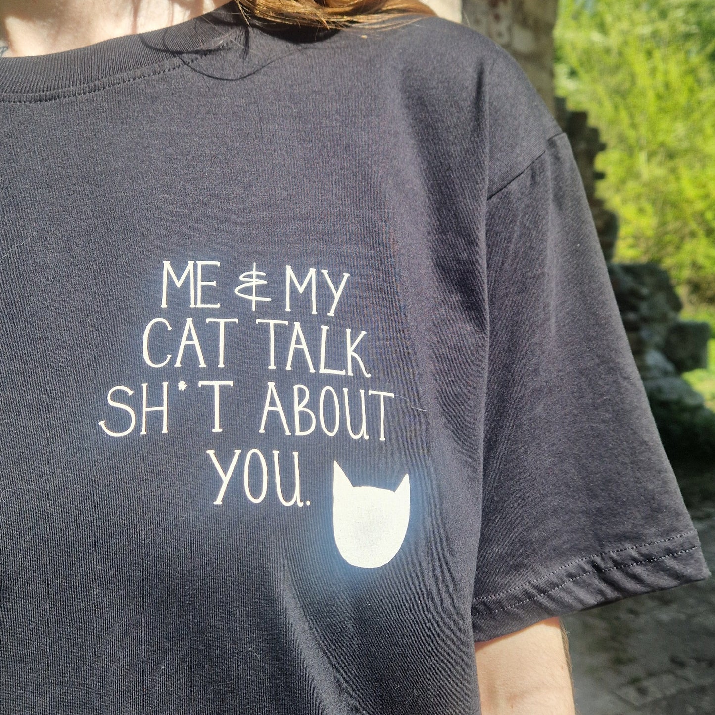 A close up of the front of the black t-shirt showing the white pocket print which reads 'ME & MY CAT TALK SH*T ABOUT YOU.' with a white cat head silhoette next to it.

