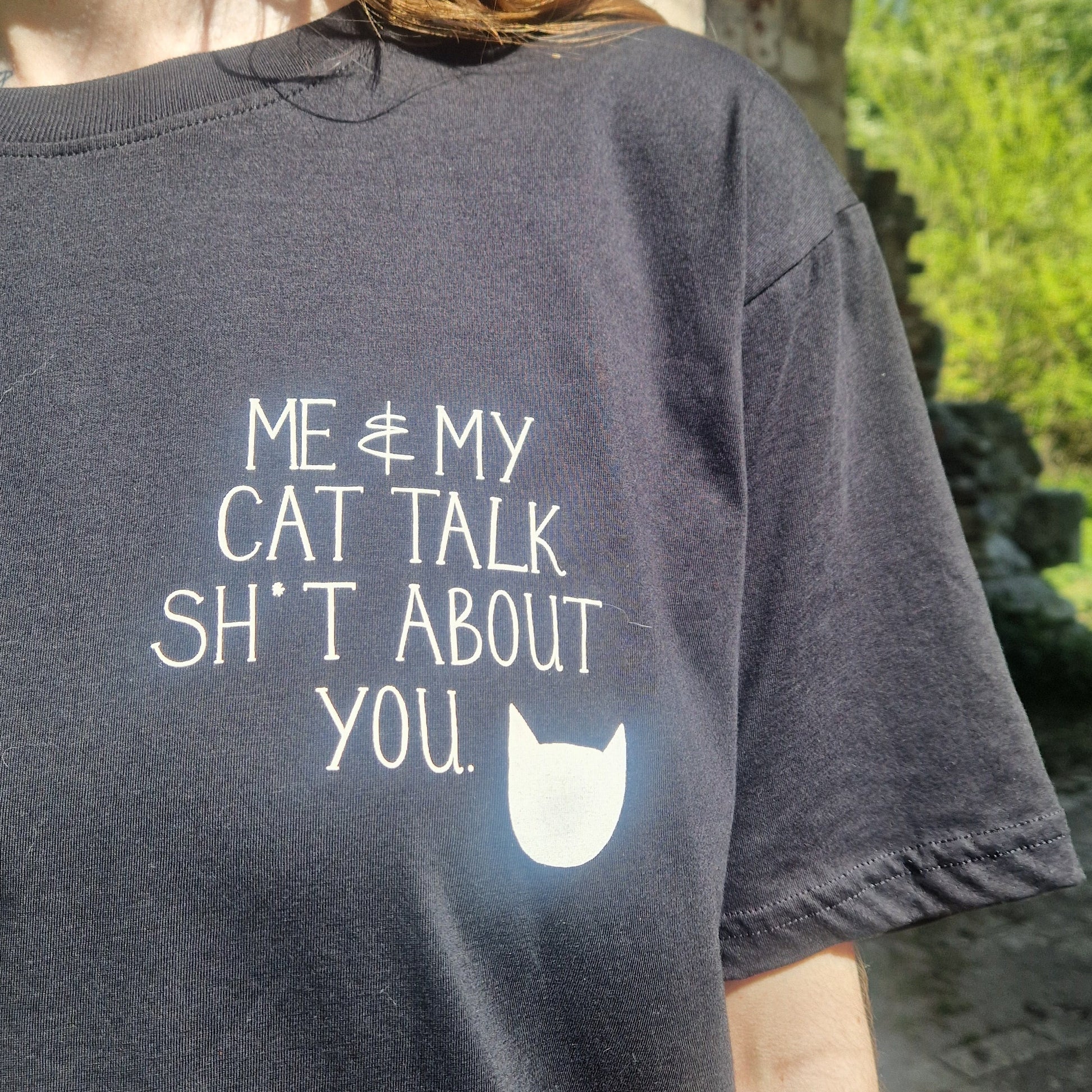 A close up of the front of the black t-shirt showing the white pocket print which reads 'ME & MY CAT TALK SH*T ABOUT YOU.' with a white cat head silhoette next to it.
