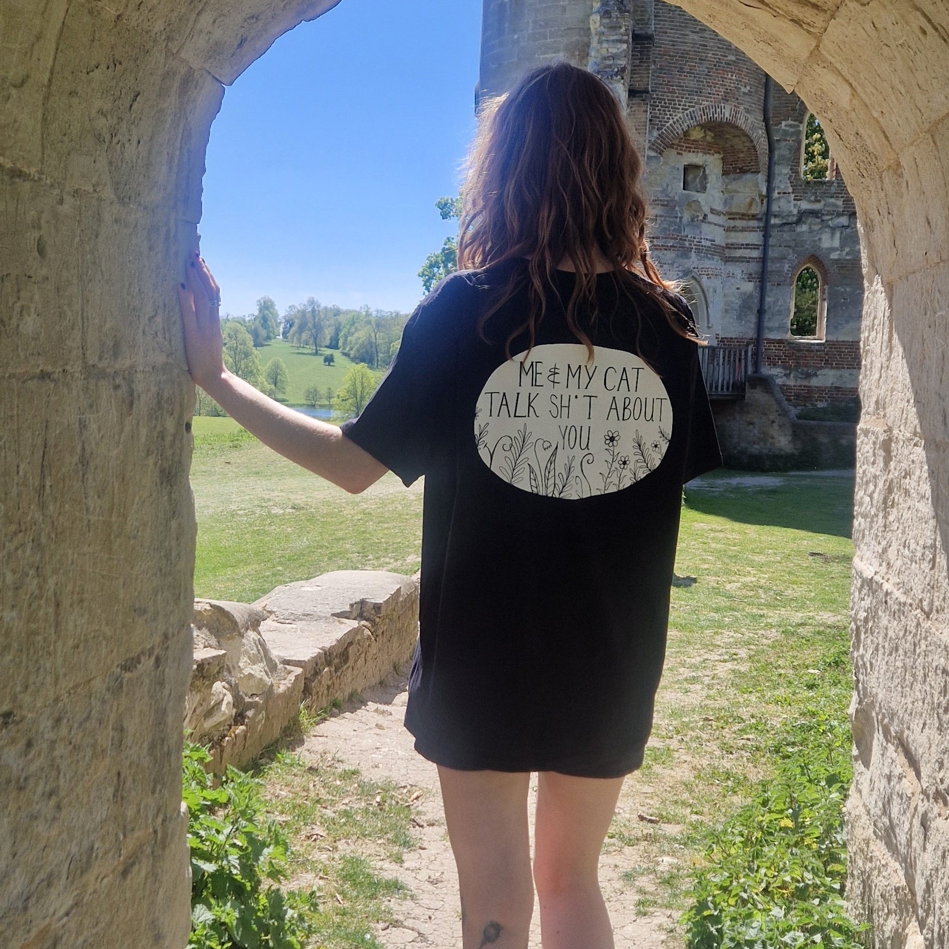 A woman in an oversized black shirt stands in an archway facing away from the camera. The back of the shirt reads 'ME & MY CAT TALK SH*T ABOUT YOU' in black in a white oval with plants growing from the bottom of it. 