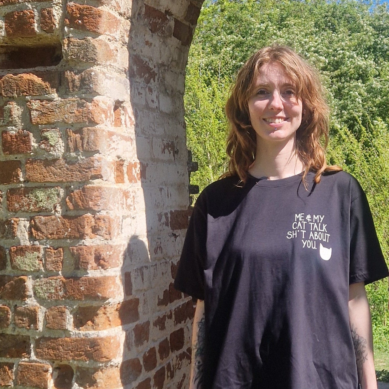 A white woman in an oversized black tshirt with a small print on the right of the shirt that reads 'ME & MY CAT TALK SH*T ABOUT YOU.' next to a white cat head silhoette. The woman stands next to an exposed brick wall. 