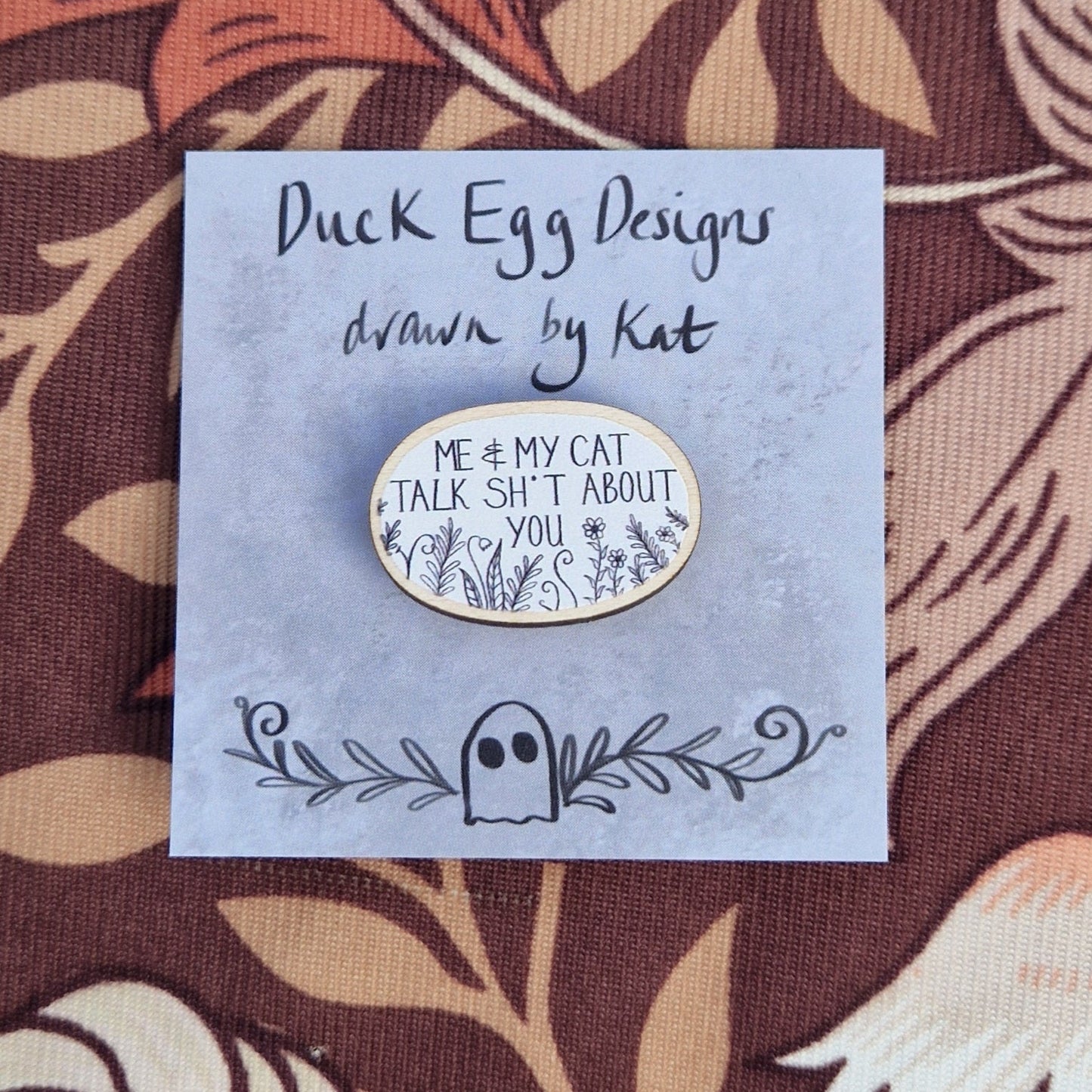 A white oval pin badge with the white words 'Me & My Cat Talk Sh*t About You' in white handwriting with black leafy fronds and flowers across the bottom. The pin sits on a grey backing card with the words 'Duck Egg Designs drawn by Kat' in black. The backing card sits on a patterned brown fabric.