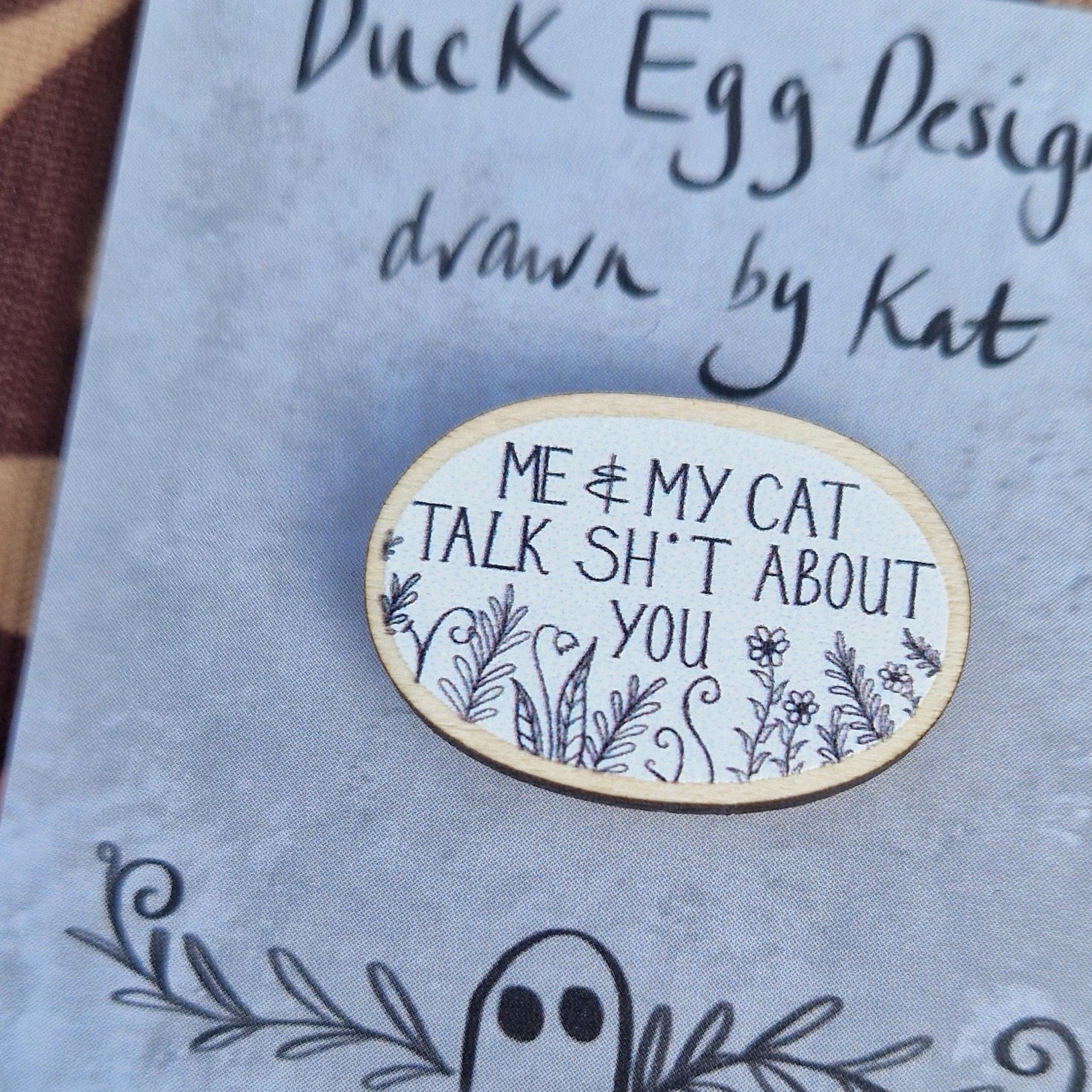 A white oval pin badge with the white words 'Me & My Cat Talk Sh*t About You' in white handwriting with black leafy fronds and flowers across the bottom. The pin sits on a grey backing card with the words 'Duck Egg Designs drawn by Kat' in black. The backing card sits on a patterned brown fabric.