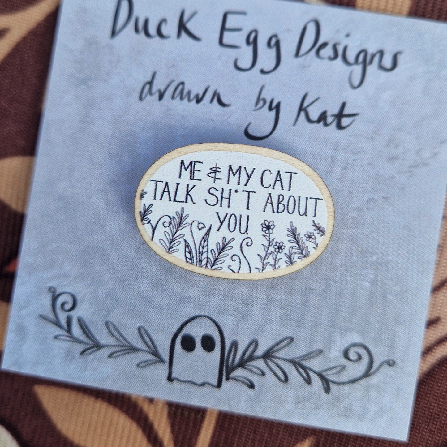 A white oval pin badge with the white words 'Me & My Cat Talk Sh*t About You' in white handwriting with black leafy fronds and flowers across the bottom. The pin sits on a grey backing card with the words 'Duck Egg Designs drawn by Kat' in black. The backing card sits on a patterned brown fabric.