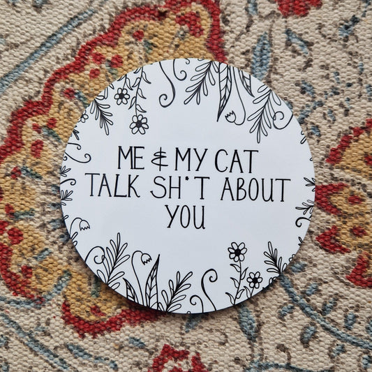 A round white coaster with a black design on it that says 'Me & My Cat Talk Sh*t About You' across the middle with plant illustrations growing in from the outside of the coaster. The coaster is on a patterned fabric background.