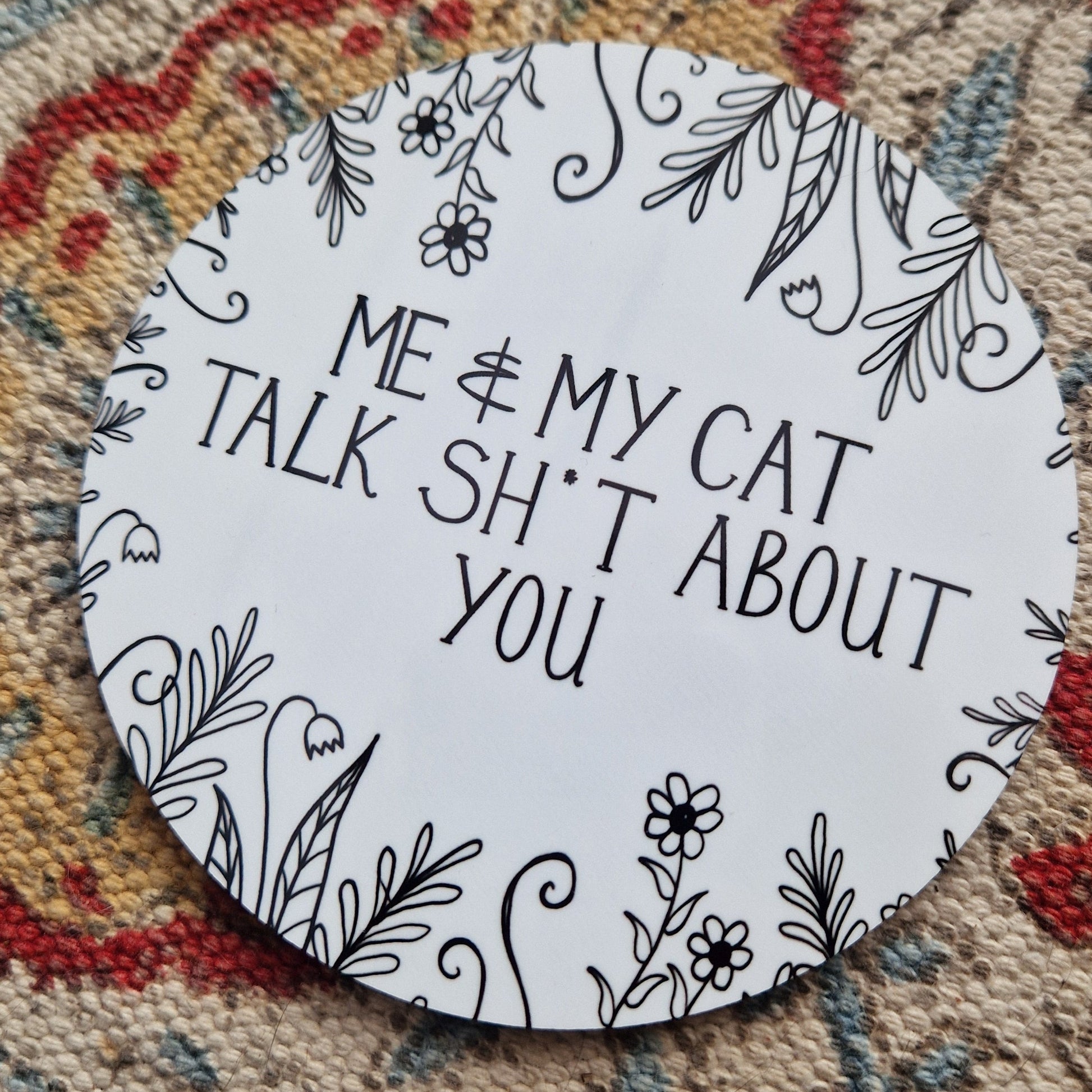 A round white coaster with a black design on it that says 'Me & My Cat Talk Sh*t About You' across the middle with plant illustrations growing in from the outside of the coaster. The coaster is on a patterned fabric background.