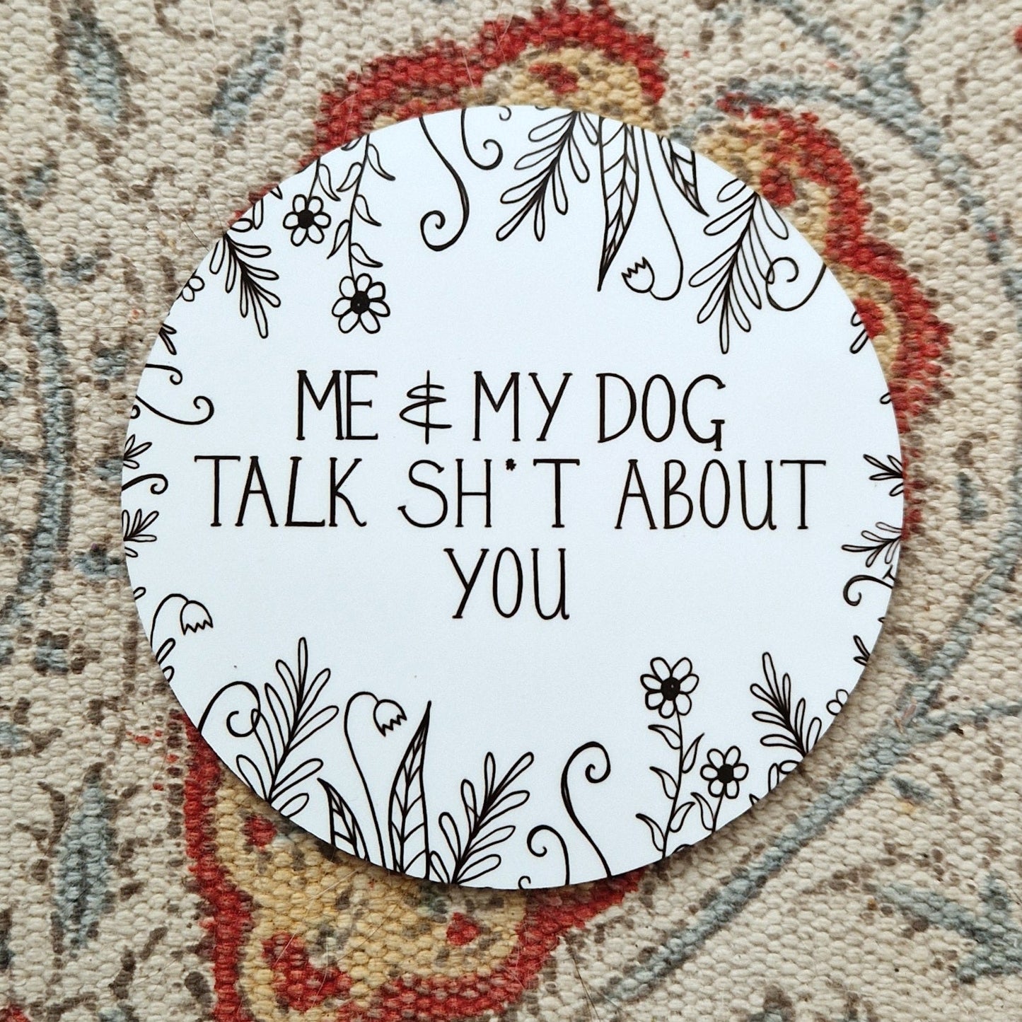 A round white coaster with the words 'Me & My Dog Talk Sh*t About You' in black handwriting across the middle and flowers and leafy vines growing in from the edges. Behind the coaster is a white patterned fabric.