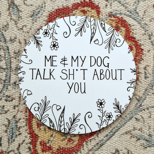 A round white coaster with the words 'Me & My Dog Talk Sh*t About You' in black handwriting across the middle and flowers and leafy vines growing in from the edges. Behind the coaster is a white patterned fabric.