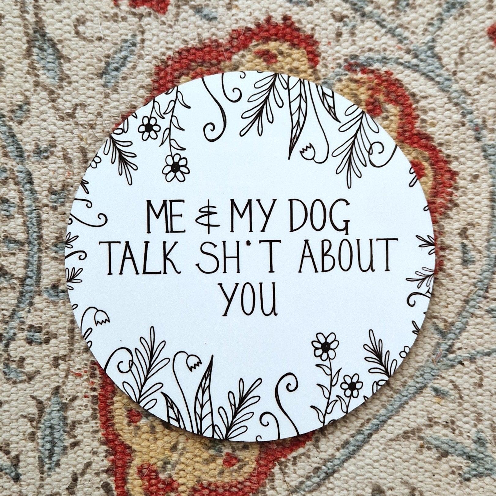 A round white coaster with the words 'Me & My Dog Talk Sh*t About You' in black handwriting across the middle and flowers and leafy vines growing in from the edges. Behind the coaster is a white patterned fabric.