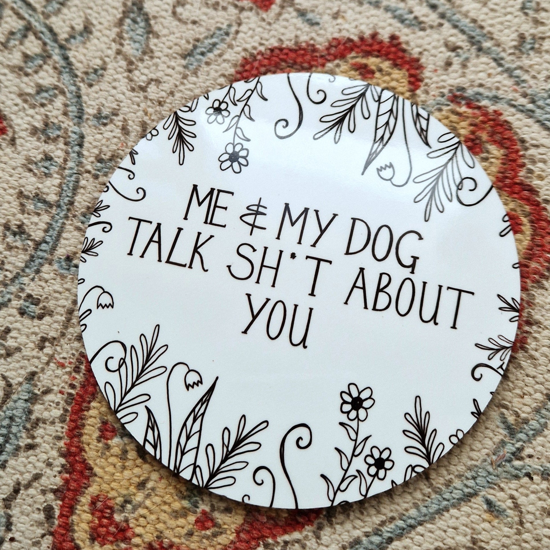 A round white coaster with the words 'Me & My Dog Talk Sh*t About You' in black handwriting across the middle and flowers and leafy vines growing in from the edges. Behind the coaster is a white patterned fabric.