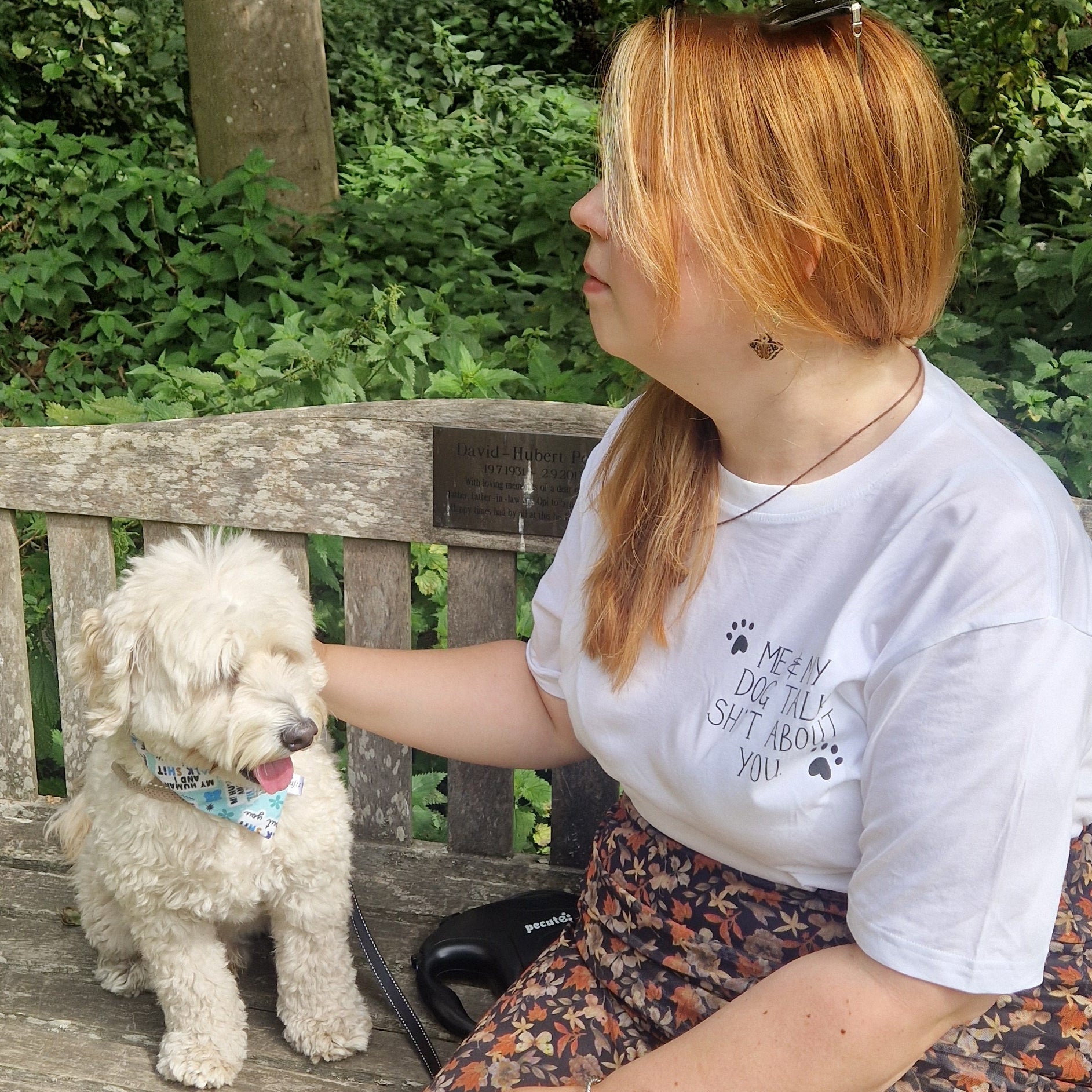 A small dog sits on the left of a wooden bench wearing a blue bandana that reads 'My Human And I Talk Shit About You' to his right is a ginger haired lady wearing a white t-shirt that reads 'Me & My Dog Talk Sh*t About You' in black with a simple pawprint design next to it.