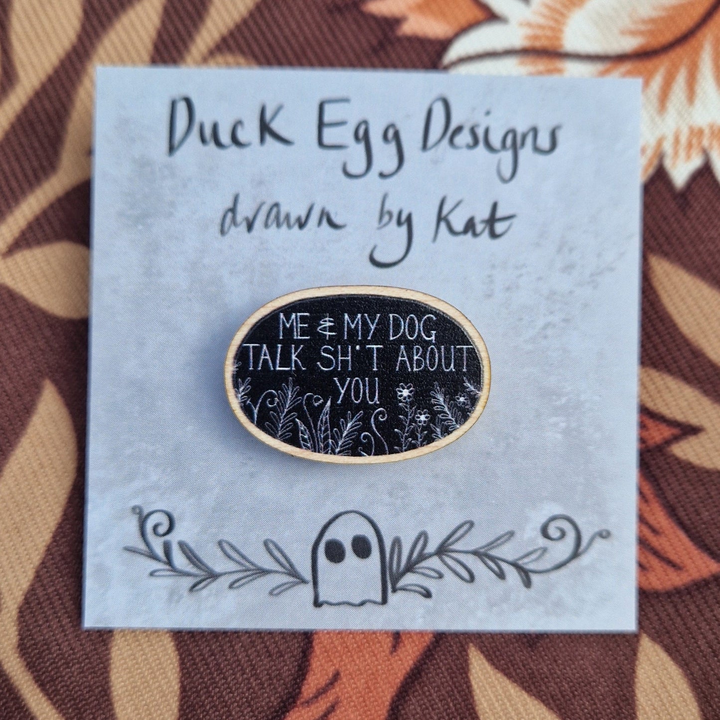 A black oval pin badge with the white words 'Me & My Dog Talk Sh*t About You' in white handwriting with white leafy fronds and flowers across the bottom. The pin sits on a grey backing card with the words 'Duck Egg Designs drawn by Kat' in black. The backing card sits on a patterned brown fabric.