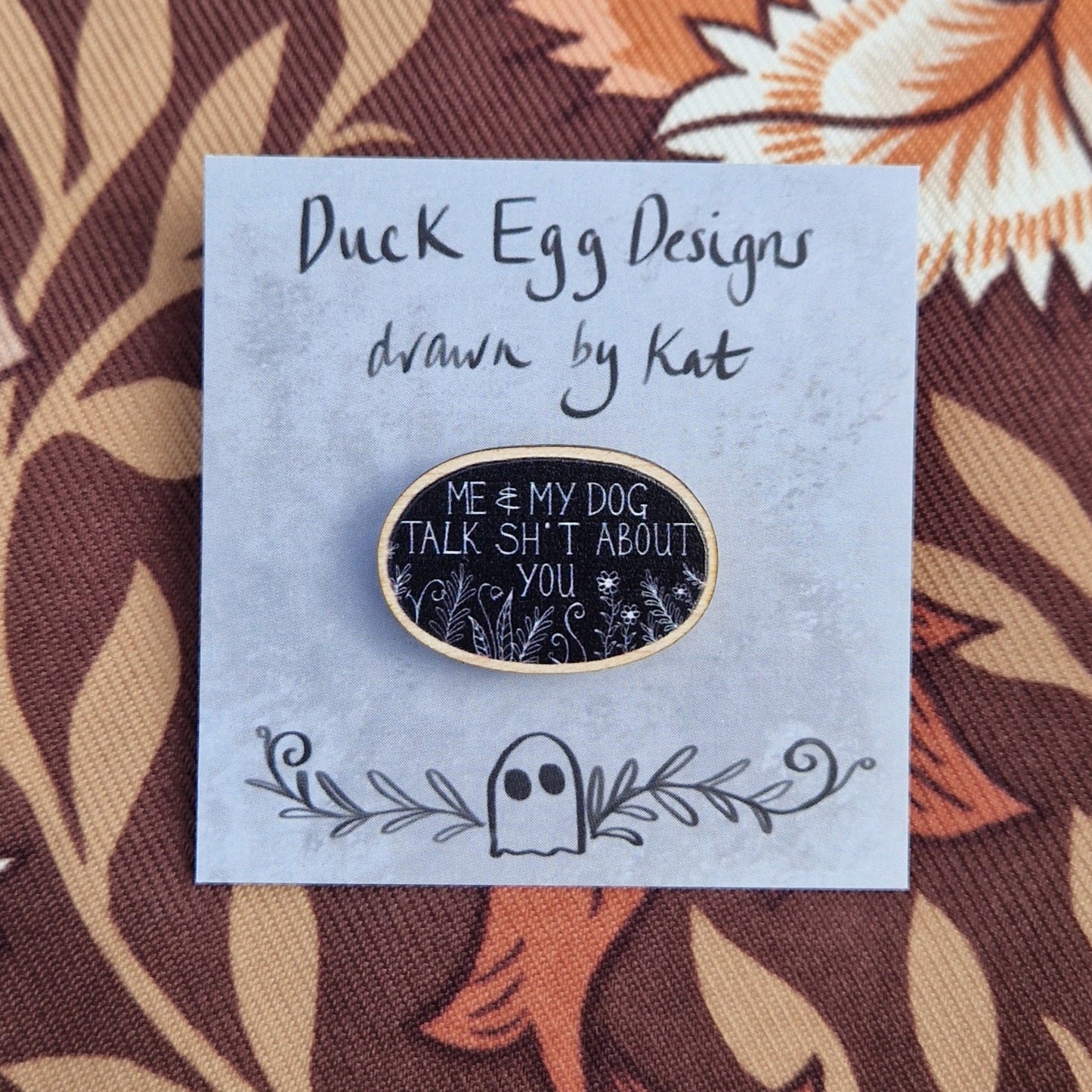 A black oval pin badge with the white words 'Me & My Dog Talk Sh*t About You' in white handwriting with white leafy fronds and flowers across the bottom. The pin sits on a grey backing card with the words 'Duck Egg Designs drawn by Kat' in black. The backing card sits on a patterned brown fabric.