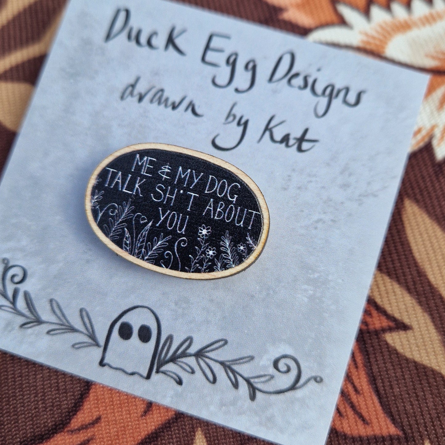 A black oval pin badge with the white words 'Me & My Dog Talk Sh*t About You' in white handwriting with white leafy fronds and flowers across the bottom. The pin sits on a grey backing card with the words 'Duck Egg Designs drawn by Kat' in black. The backing card sits on a patterned brown fabric.