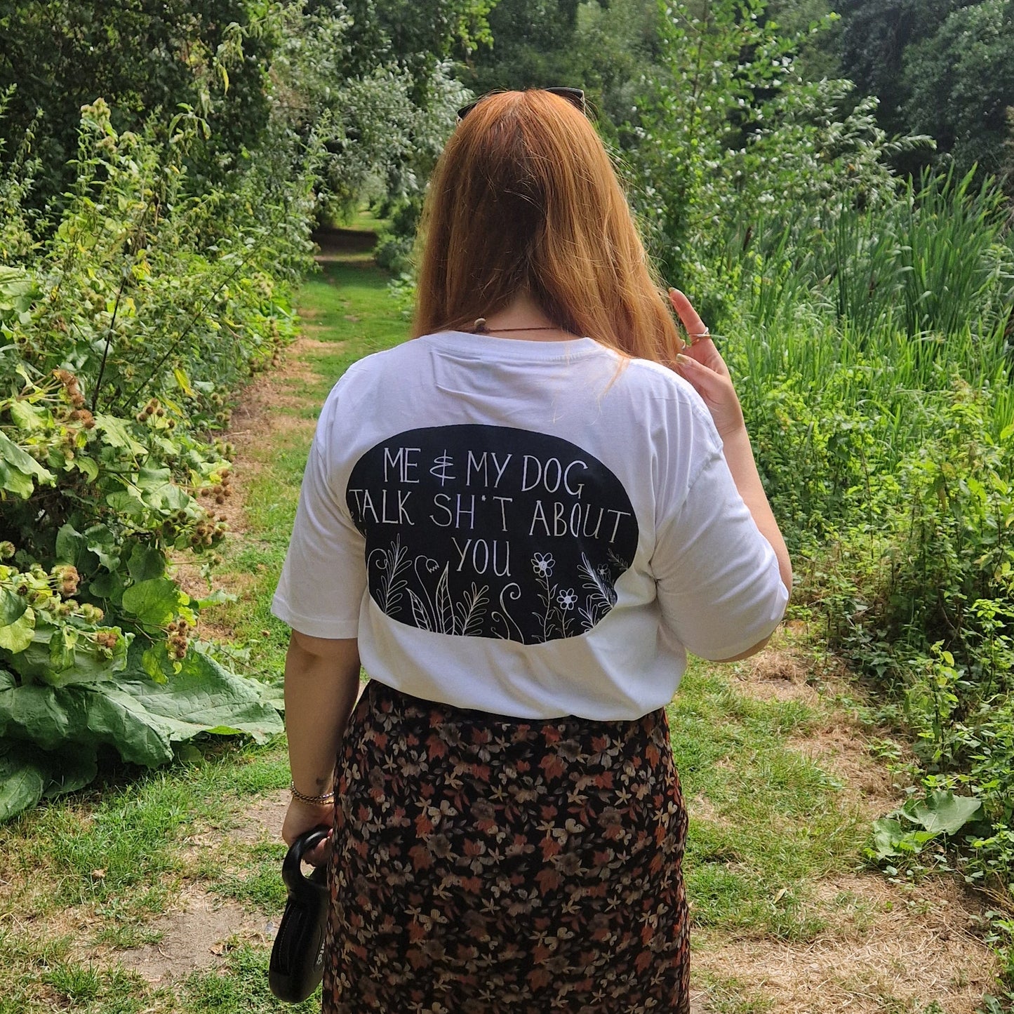 A lady with long ginger hair and sunglasses on her head faces away from the camera showing off the back of her white t-shirt that reads 'Me & My Dog Talk Sh*t About You" in white within a black oval with flowers and leafy fronds growing from the bottom. She wears a n autumnal orange and black skirt with it and has a black dog lead in her left hand. Behind her is a green grassy path with trees across the edges.