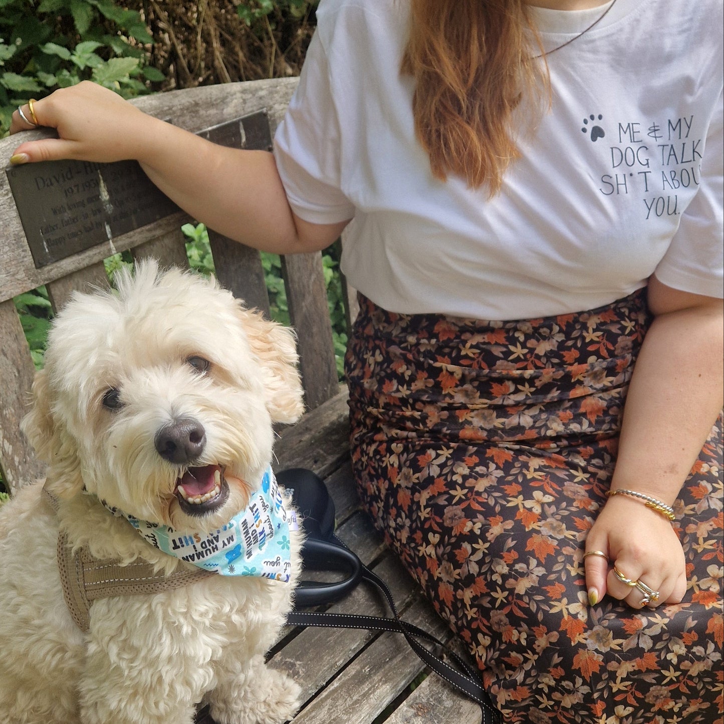 A small dog sits on the left of a wooden bench wearing a blue bandana that reads 'My Human And I Talk Shit About You' to his right is a ginger haired lady wearing a white t-shirt that reads 'Me & My Dog Talk Sh*t About You' in black with a simple pawprint design next to it.