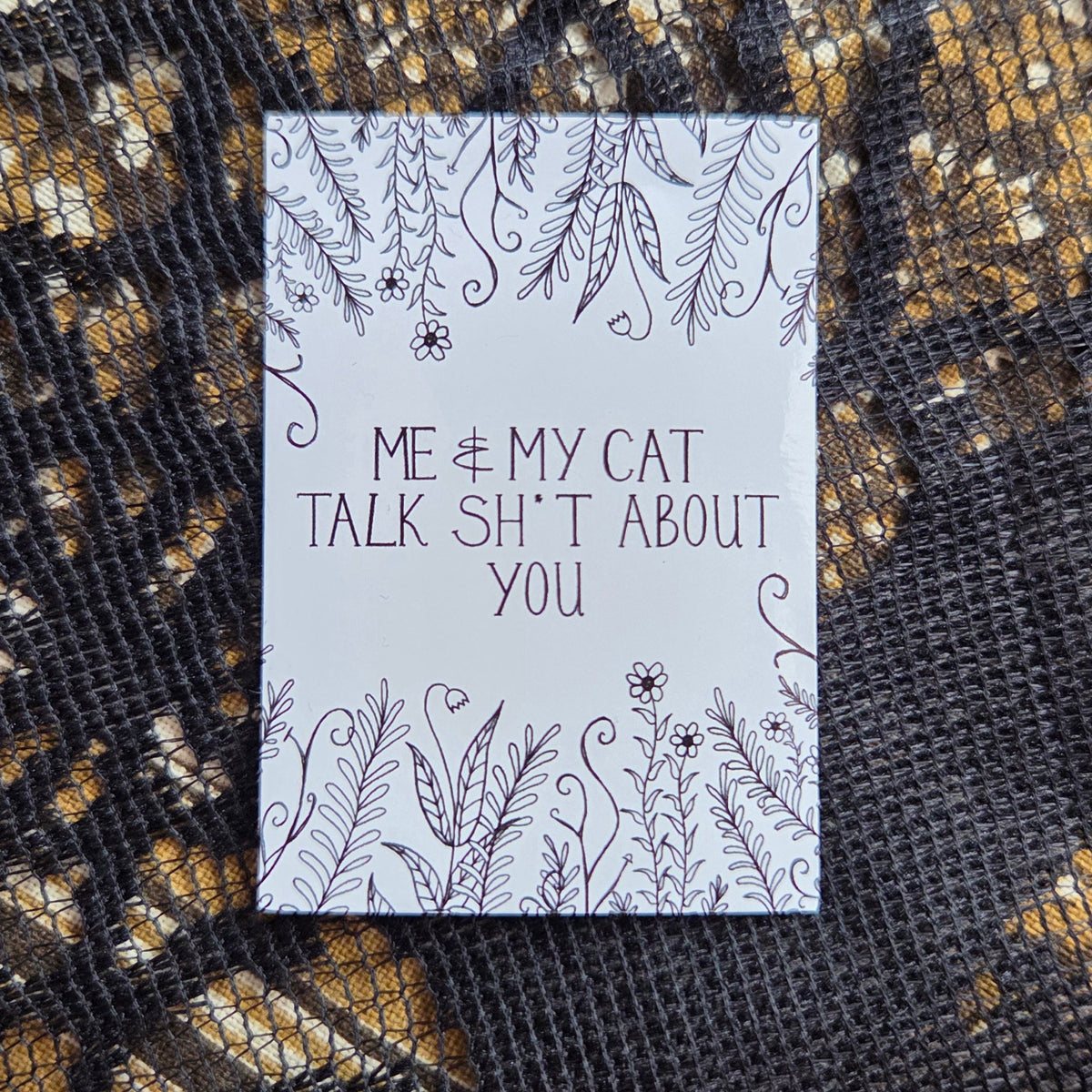 A white rectangular sticker with the words 'Me & my cat talk shit about you' in black handwriting across the middle, with leafy vines and flowers growing from the top and bottom of the design. Behind the sticker is a brown floral fabric with a black lace overlay.