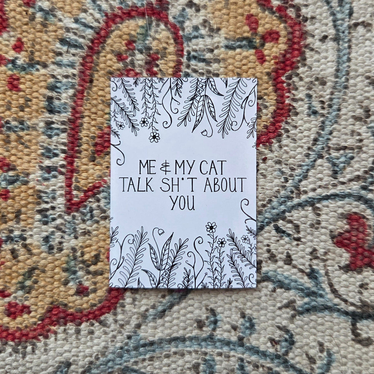 A white rectangular sticker with the words 'Me & my cat talk shit about you' in black handwriting across the middle, with leafy vines and flowers growing from the top and bottom of the design. Behind the sticker is a colourful patterned fabric.