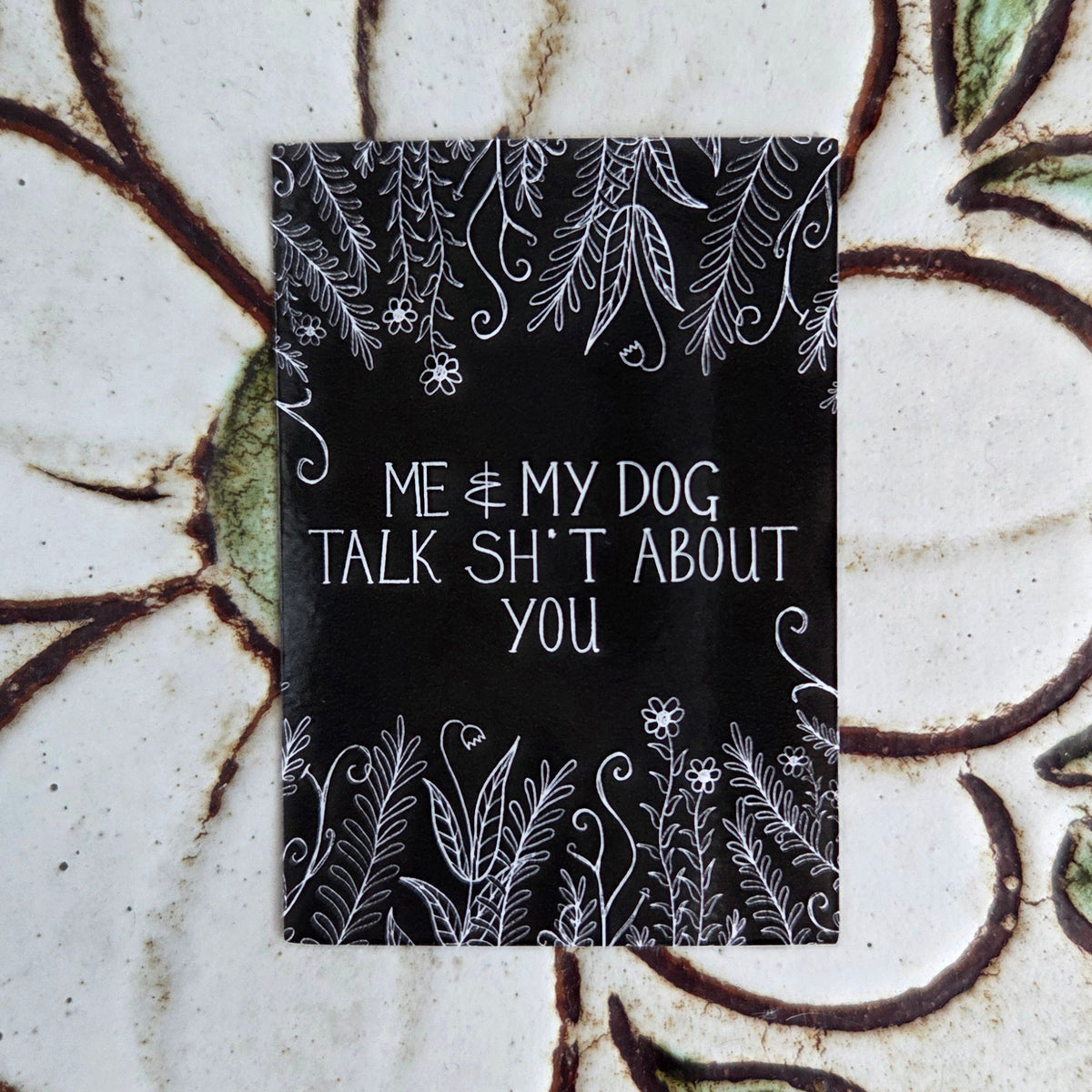 A black rectangular sticker with the words 'Me & my dog talk shit about you' in white handwriting across the middle, with leafy vines and flowers growing from the top and bottom of the design. Behind the sticker is a floral tile background.
