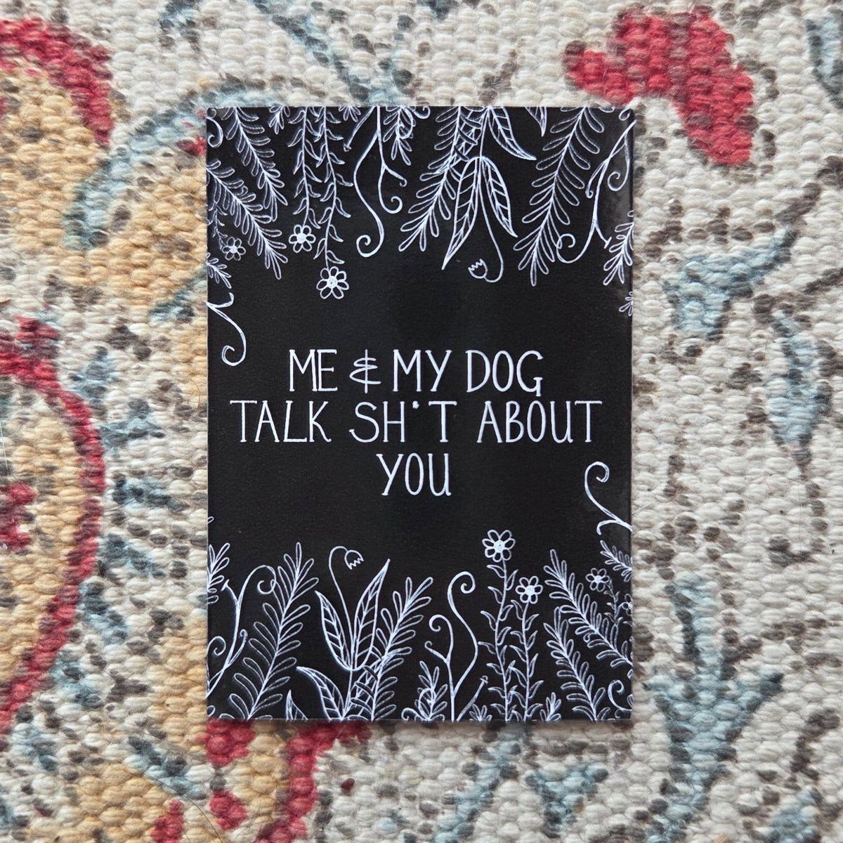 A black rectangular sticker with the words 'Me & my dog talk shit about you' in white handwriting across the middle, with leafy vines and flowers growing from the top and bottom of the design. Behind the sticker is a colourful fabric background.