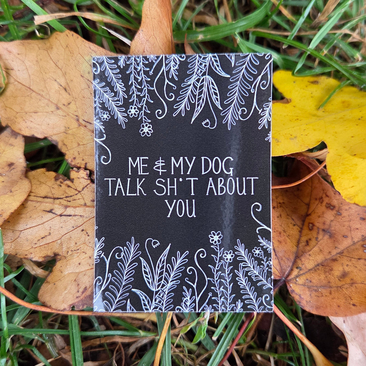A black rectangular sticker with the words 'Me & my dog talk shit about you' in white handwriting across the middle, with leafy vines and flowers growing from the top and bottom of the design. Behind the sticker grassy background with autumnal yellow and brown leaves.