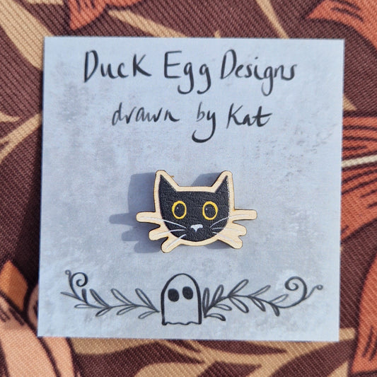 A small black cat head pin badge sits on a grey backing card with the words 'Duck Egg Designs drawn by Kat' across the top. The cat has yellow eyes and a grey nose and whiskers. Behind the backing card is a brown patterned fabric.