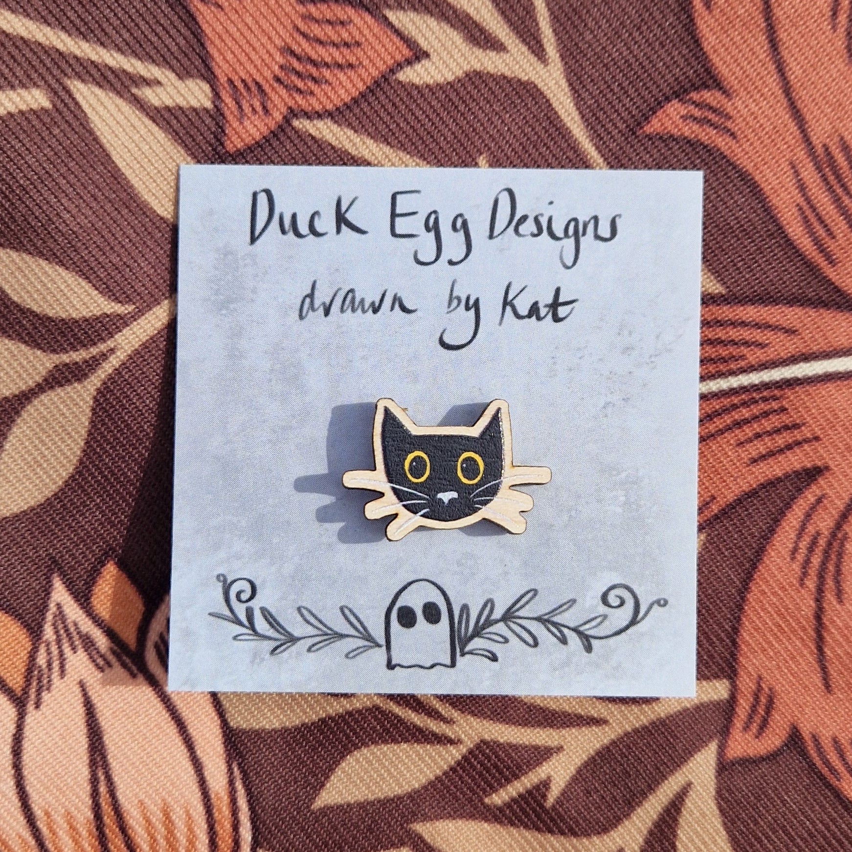 A small black cat head pin badge sits on a grey backing card with the words 'Duck Egg Designs drawn by Kat' across the top. The cat has yellow eyes and a grey nose and whiskers. Behind the backing card is a brown patterned fabric.