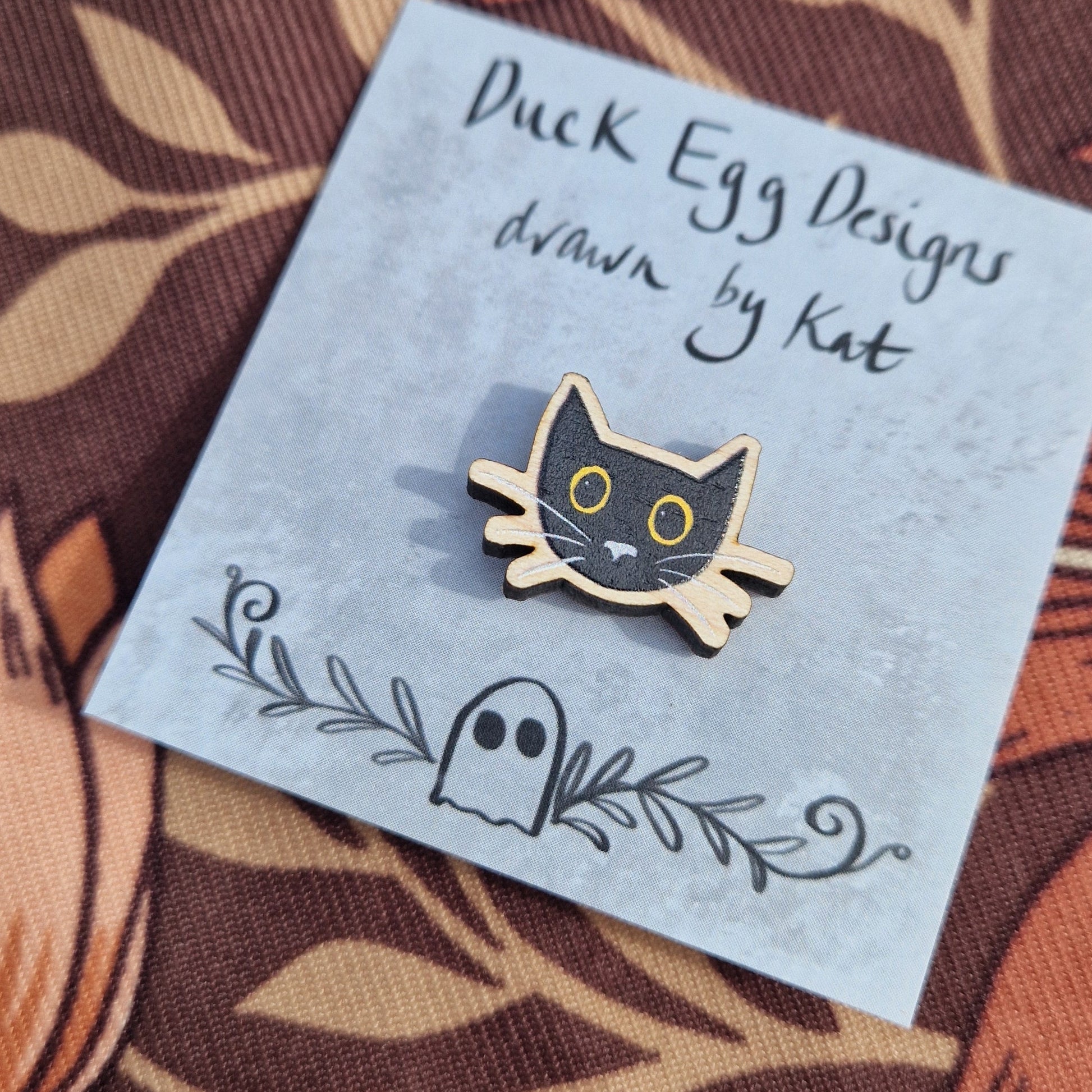 A small black cat head pin badge sits on a grey backing card with the words 'Duck Egg Designs drawn by Kat' across the top. The cat has yellow eyes and a grey nose and whiskers. Behind the backing card is a brown patterned fabric.