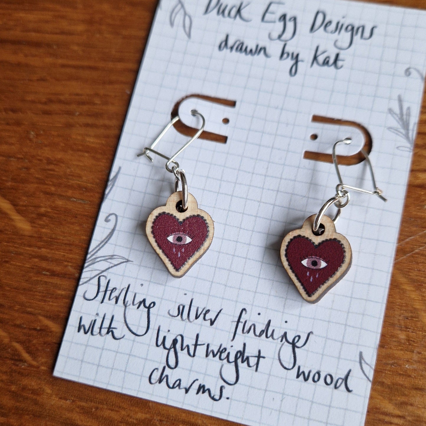 A close up of the red crying heart earring charms on sterling silver hooks. The earrings sit on a white backing card which features the words 'Sterling silver findings with lightweight wood charms'. Behind the earrings is a wooden tabletop.