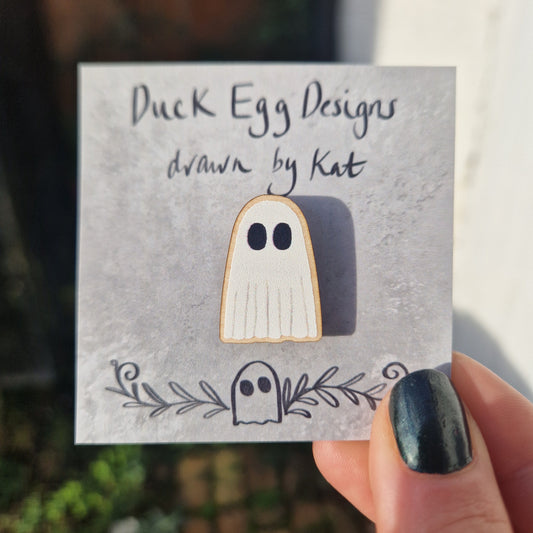 A wooden white ghost pin badge sits on a mottled grey backing card featuring the words 'Duck Egg Designs drawn by Kat'. The backing card is held by a white hand with black nail polish in front of an overgrown garden.