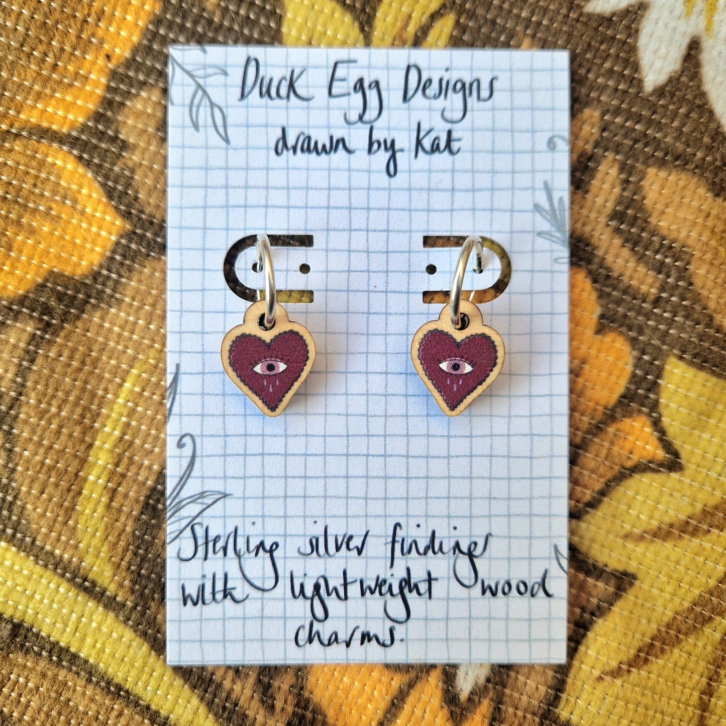 Mini crying heart charms in deep red with a dusky pink eye hanging from mini sterling silver hoops on a white backing card. Behind them is a retro floral fabric in oranges and browns.