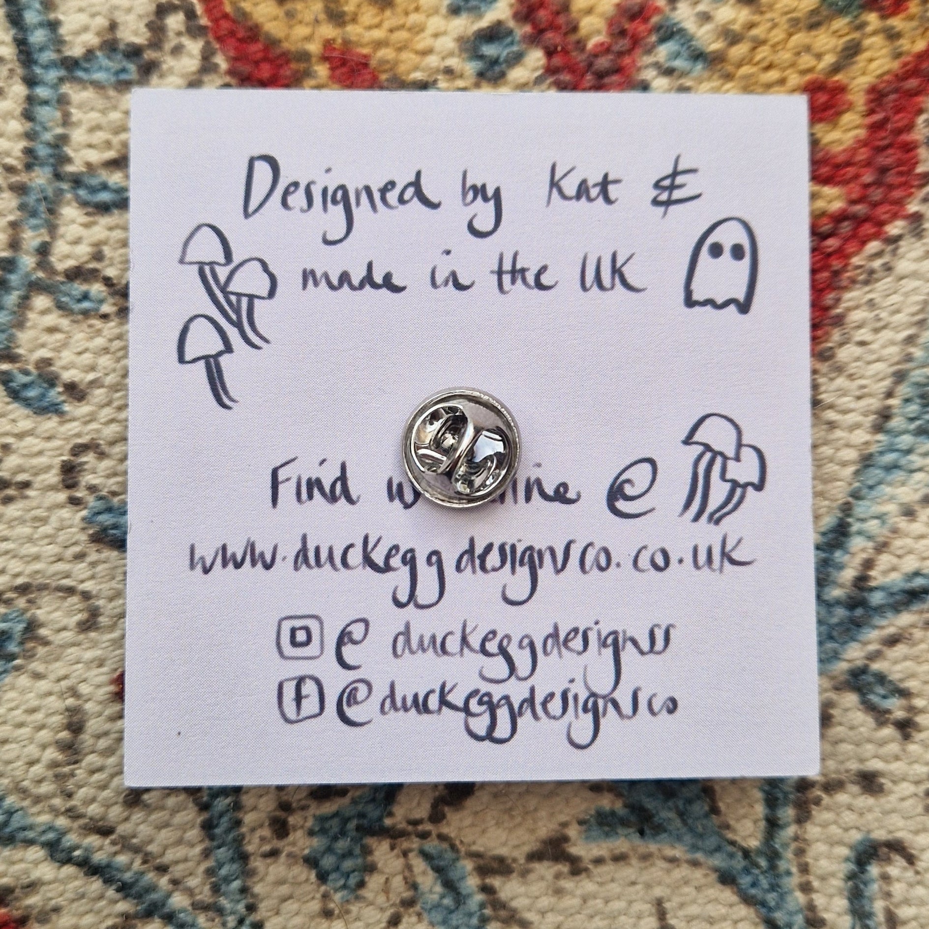 The back of the pin badge on the backing card showing the silver metal butterfly clutch back as well as the words 'Designed by Kat & made in the UK' alongside some simple fungi and ghost illustrations. Behind the backing card you can see a patterned fabric background.