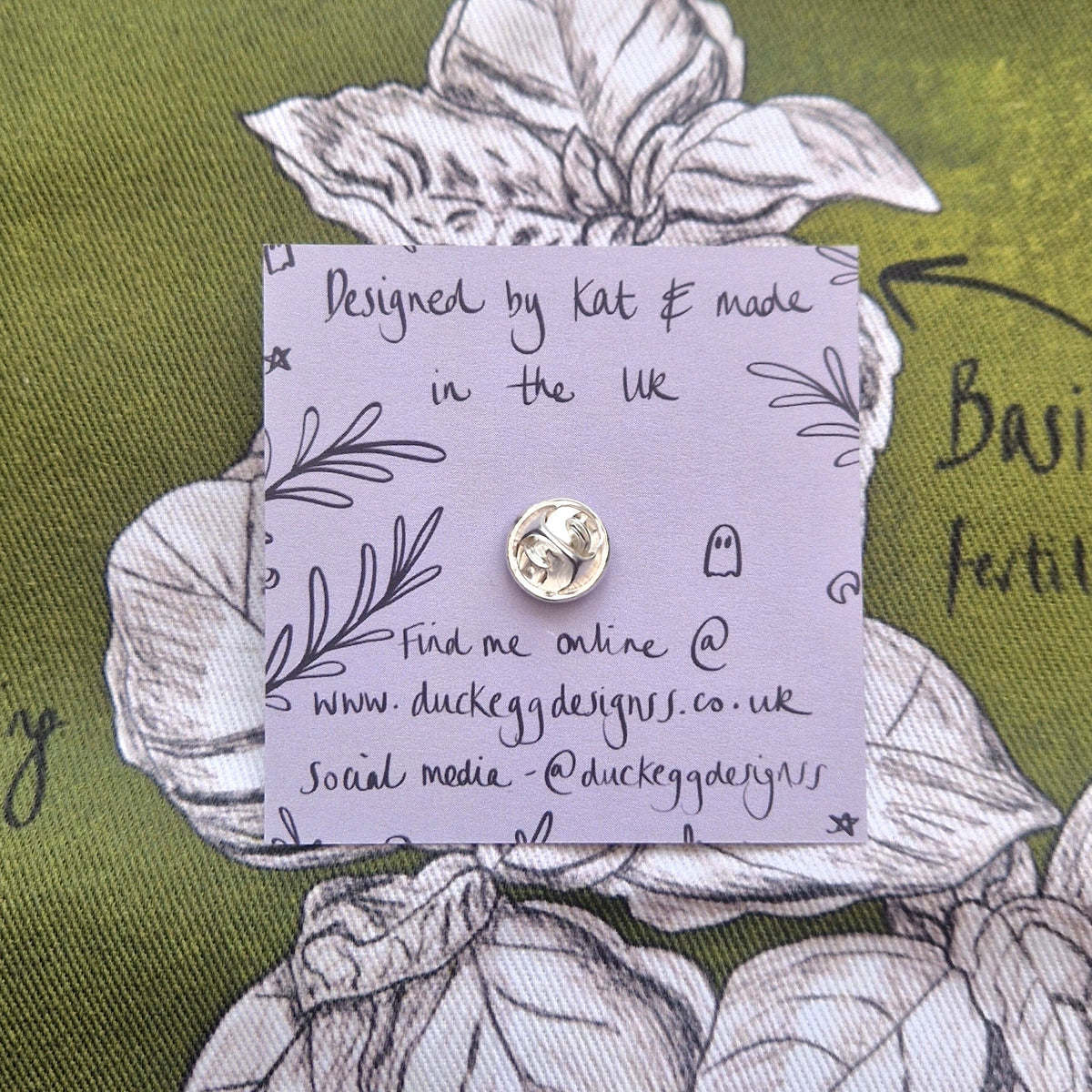 The back of the pin badge, showing the silver coloured butterfly clasp as well as the light purple back of the backing card. Behind the backing card is a rich green fabric with a black and white plant design.