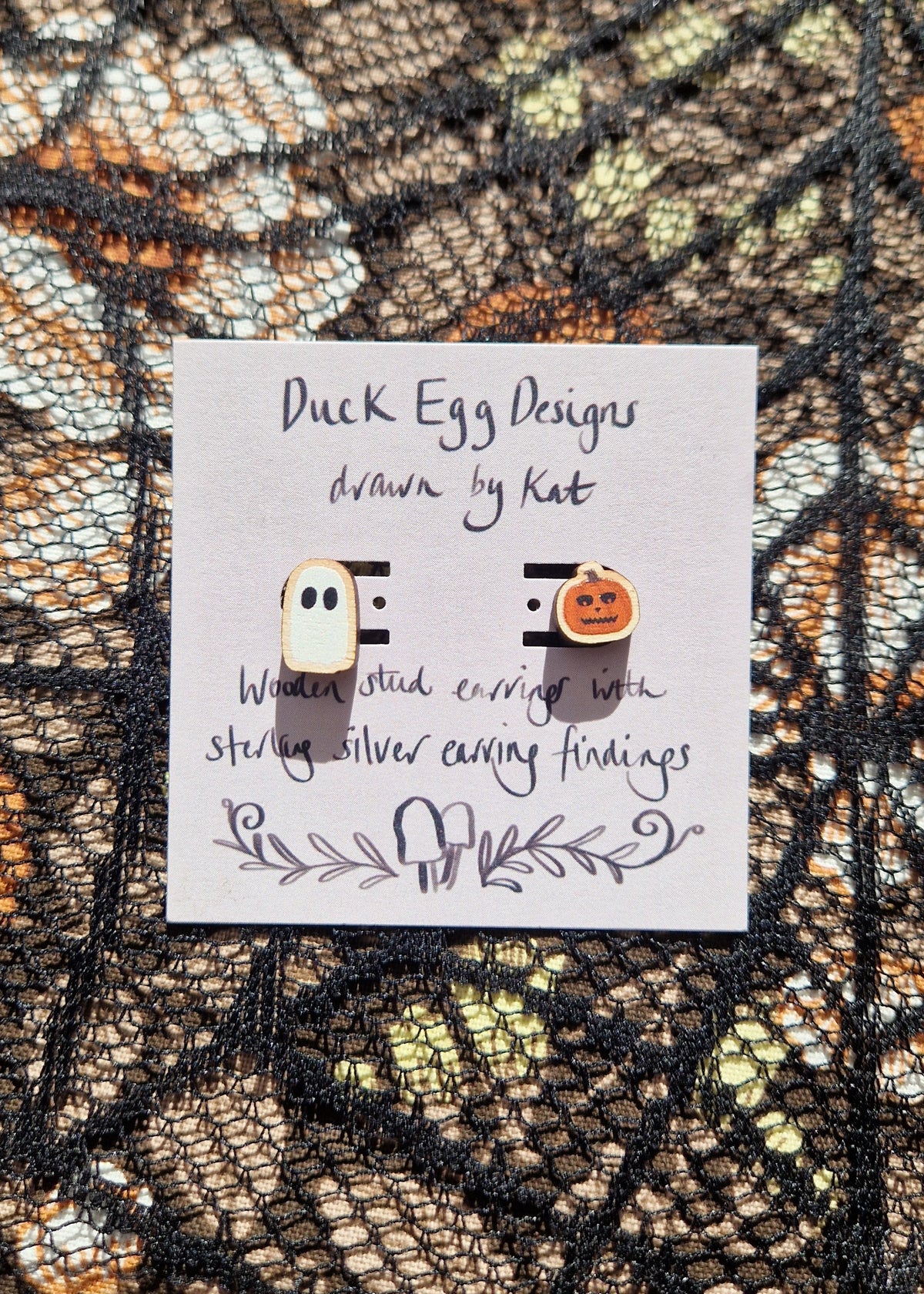 A mismatched pair of earrings with one ghost and one carved pumpkin stud sit on a light purple backing card with the words 'Duck Egg Designs drawn by Kat' in black writing. Behind the backing card is a black lace and floral fabric background.
