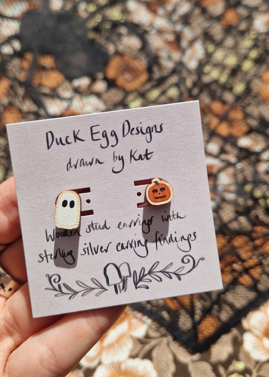 A mismatched pair of earrings with one ghost and one carved pumpkin stud sit on a light purple backing card with the words 'Duck Egg Designs drawn by Kat' in black writing. The earrings are held on a white hand above a black lace and floral fabric background.