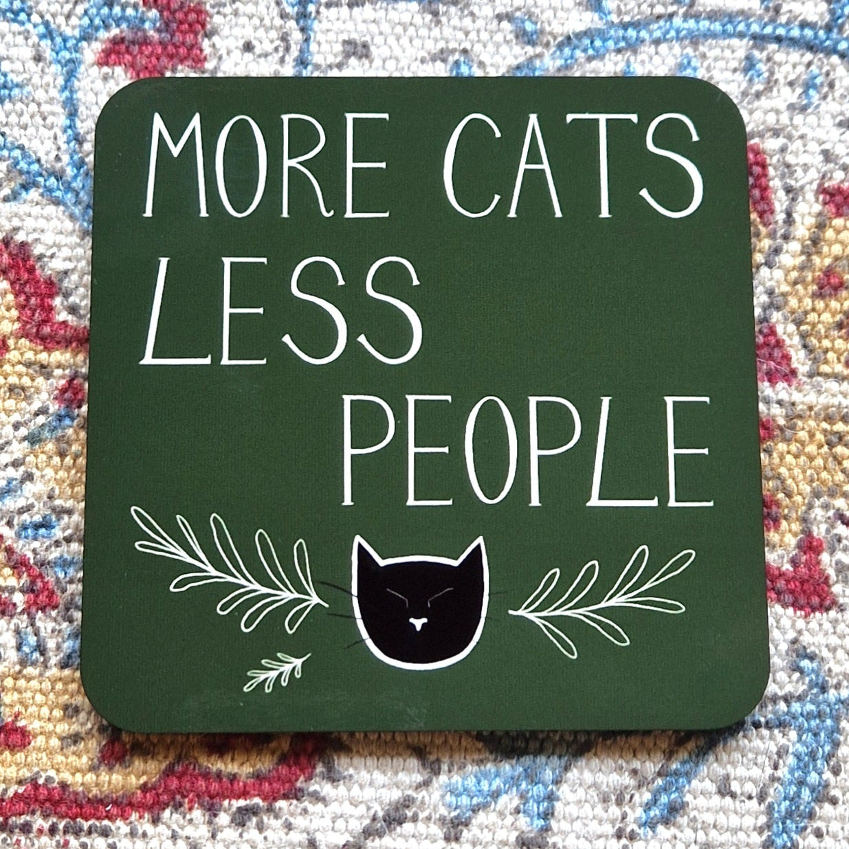 A square mossy green coaster featuring the words 'MORE CATS LESS PEOPLE' in white handwriting above a black cat head and leafy tendril design.